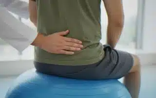 Posture treatment specialist assessing a patient’s lower back alignment while they sit on an exercise ball to improve posture, mobility, and overall well-being.